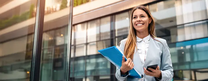 Businesswoman smiling, holding a file and phone outside a modern office building.